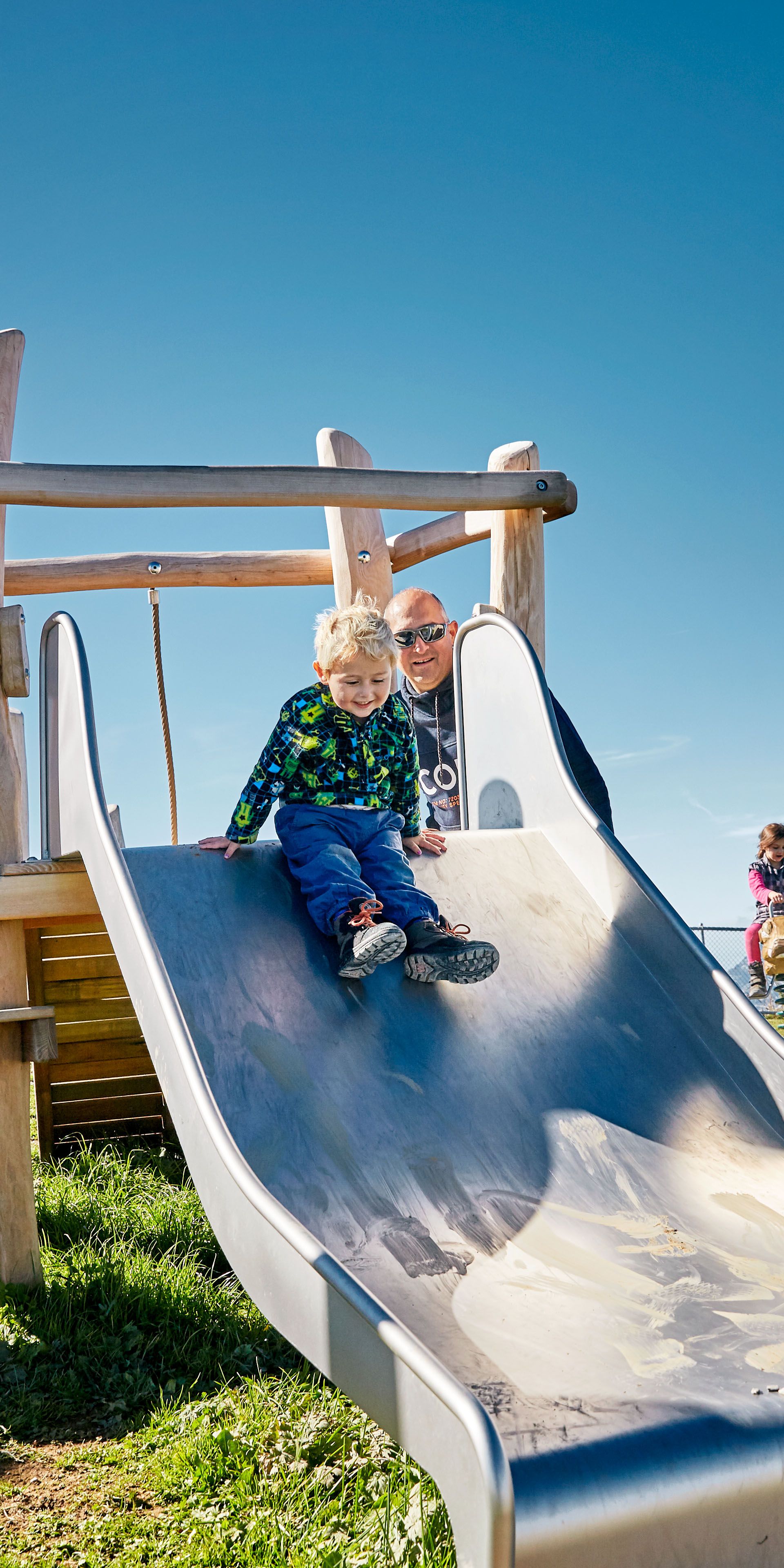Kind rutscht auf einer Rutschbahn auf einem Spielplatz in alpiner Umgebung.
